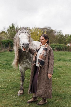 Grey dappled horse shows playful expression while standing with person in brown winter coat in grassy field.