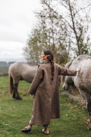 A figure wearing a long brown coat stands beside a dappled grey horse in a pastoral autumn field.