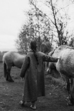 A person wearing a long winter coat stands between two horses in a black and white outdoor setting.