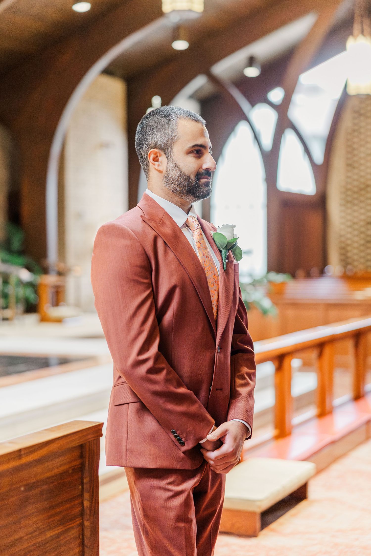 A person wearing a brown suit stands thoughtfully in a church interior with wooden pews and arched windows.