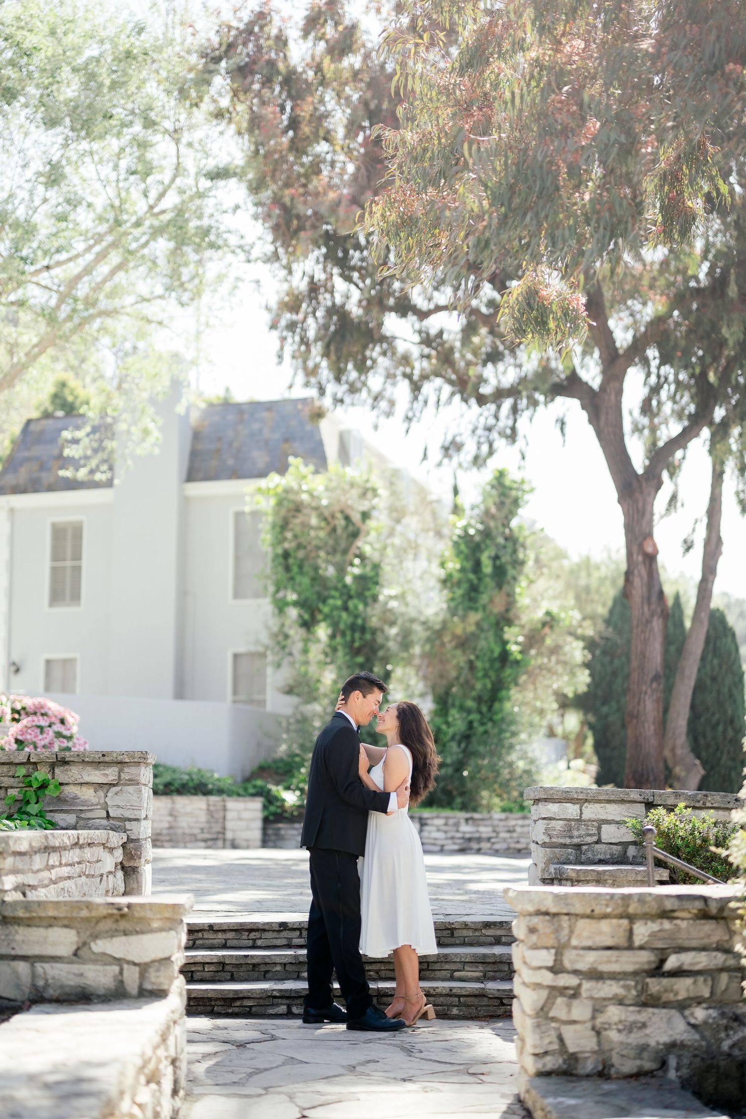 A romantic engagement photo session captured in a serene garden setting with stone walls and trees in the background.