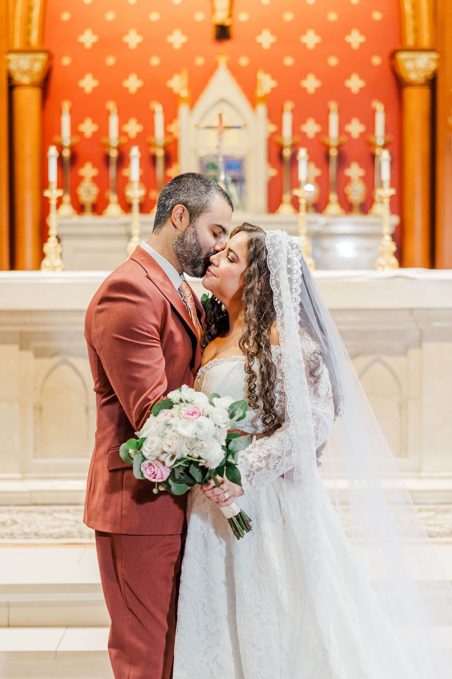 A couple shares an intimate moment at the altar of a church with red walls and candlesticks, the bride holding a white bouquet.