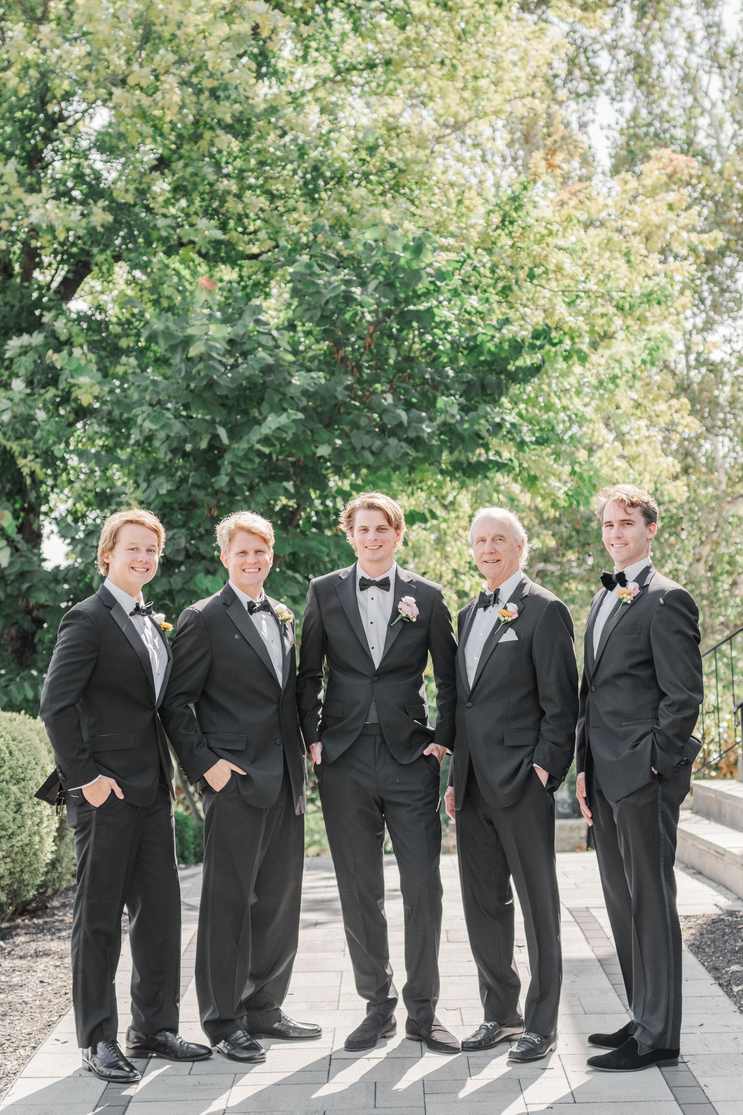 Groomsmen in black tuxedos lined up for formal photos on a stone pathway.