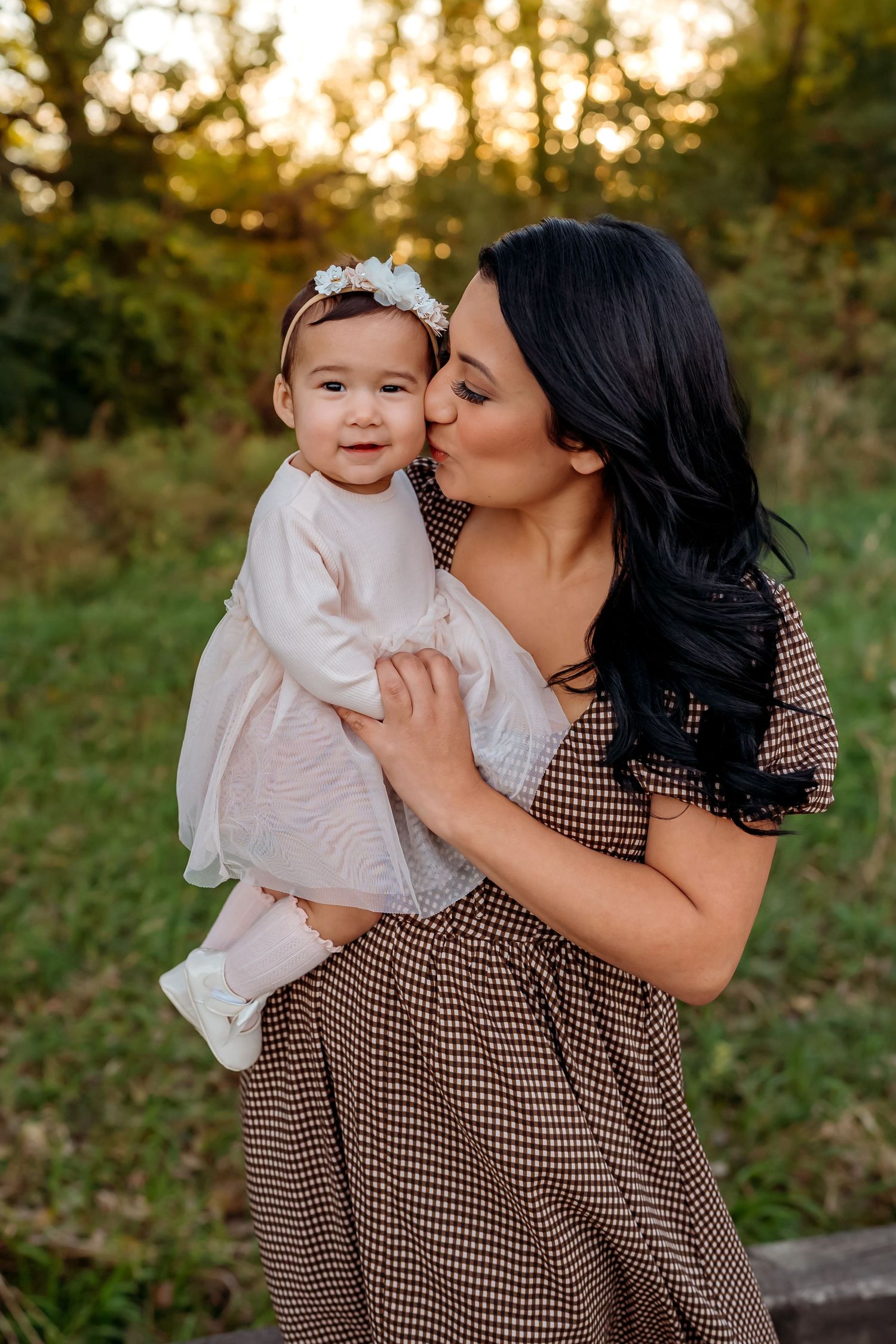 A tender moment between parent and baby in white dress against a golden sunset backdrop in an outdoor setting.