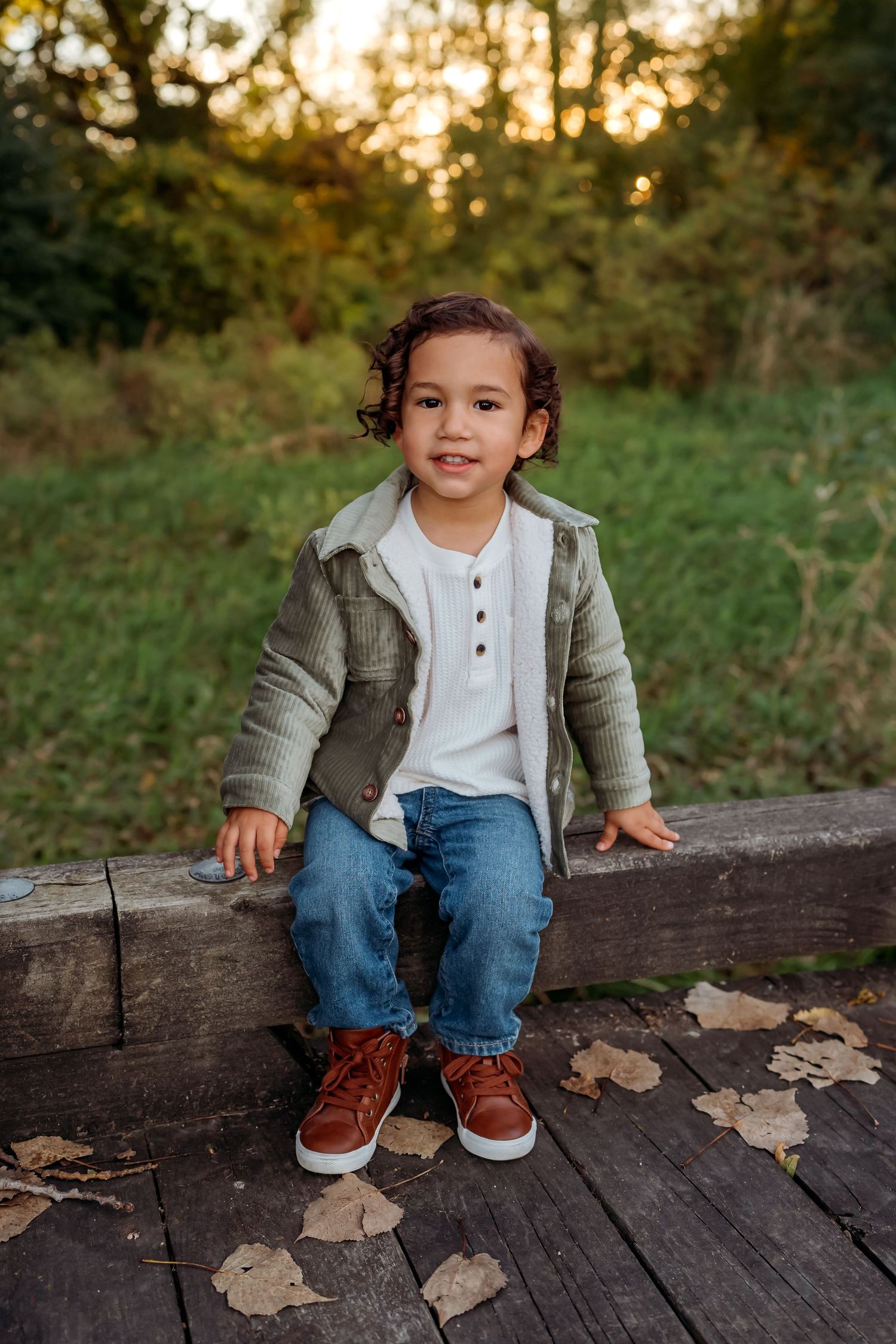 A child sits on a wooden log surrounded by autumn leaves at sunset wearing a gray jacket and jeans.