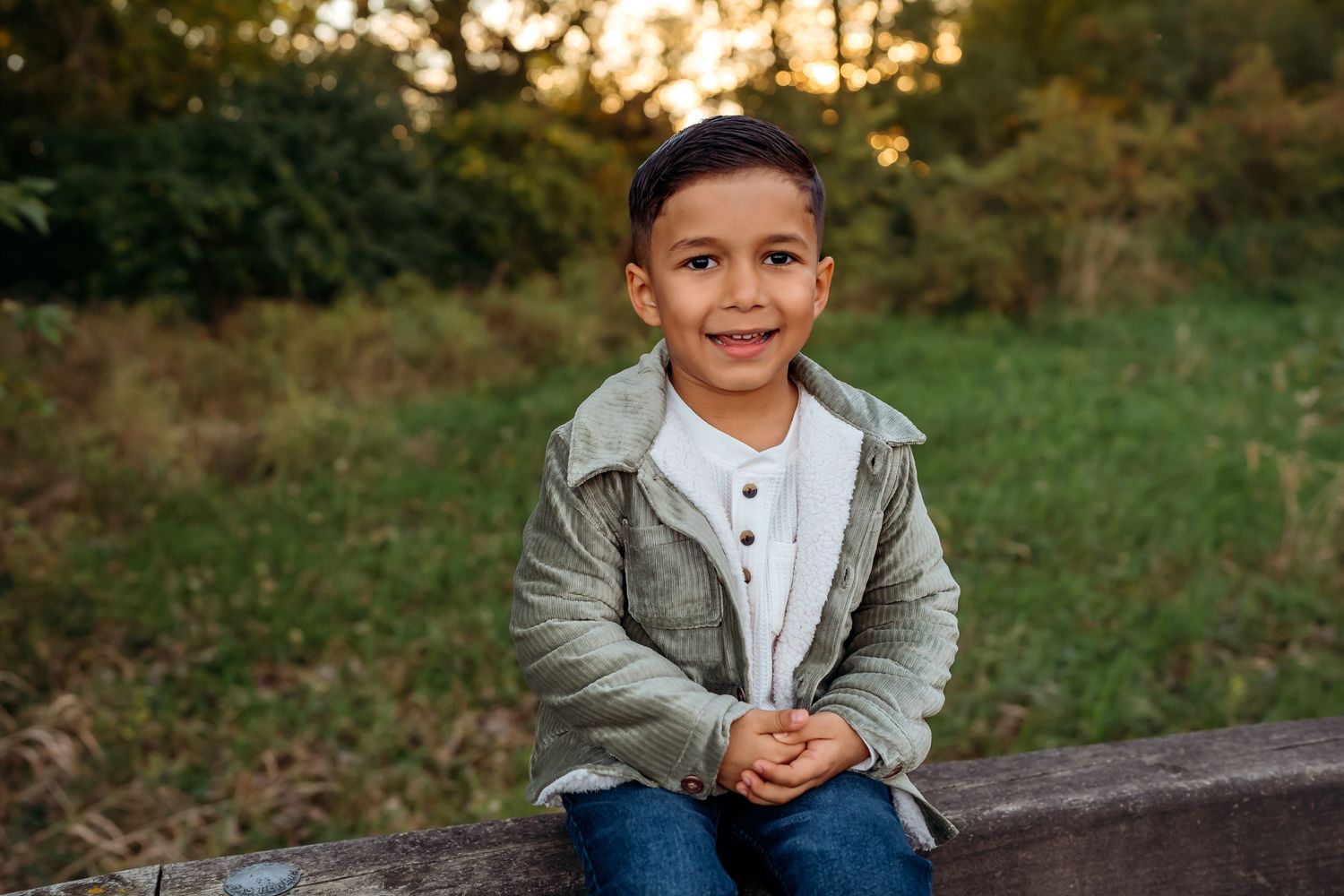 Young child wearing a gray jacket and jeans sits outdoors at sunset with a warm smile.