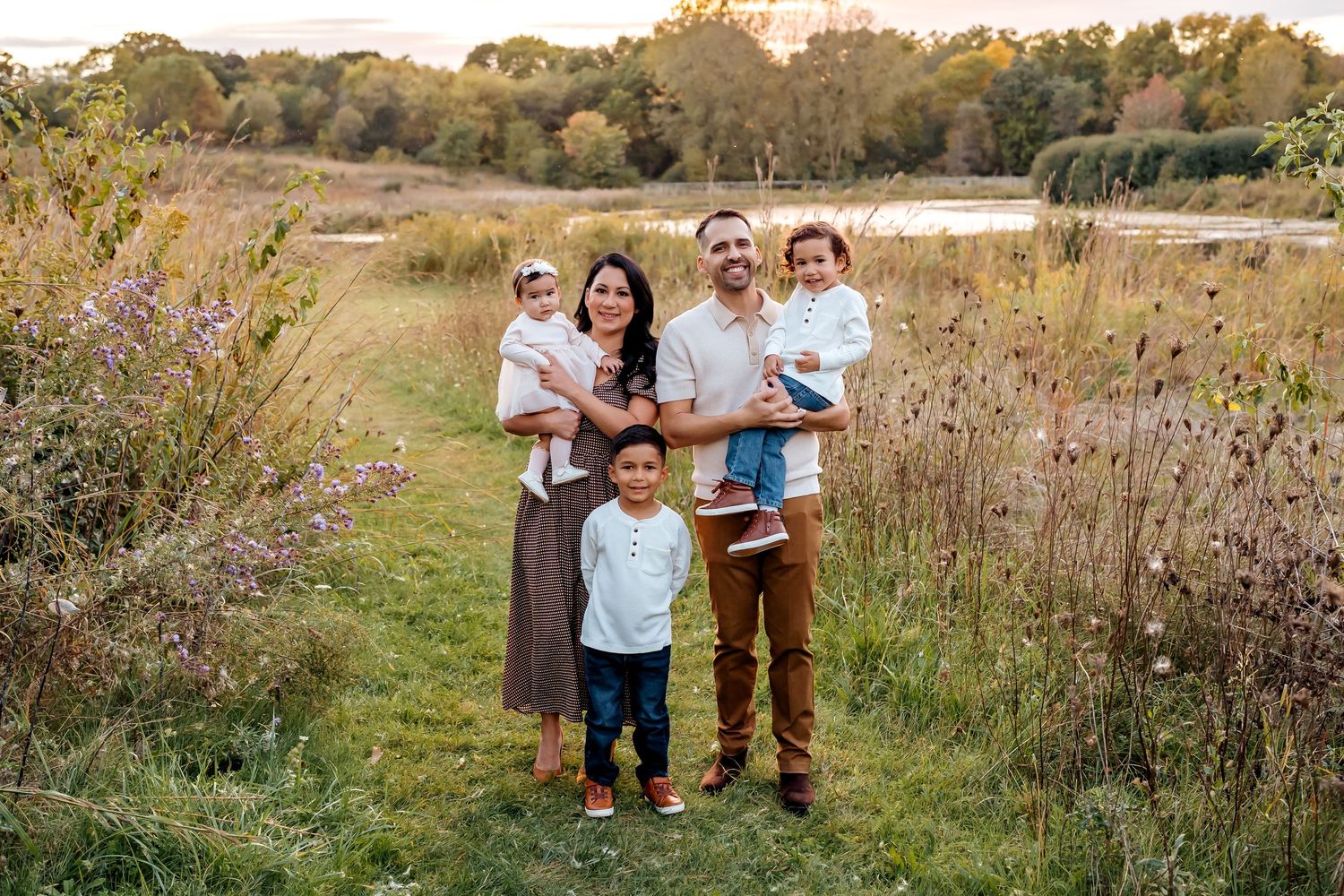 A family enjoys a candid photoshoot together in a scenic autumn meadow with tall grass and wildflowers at sunset.