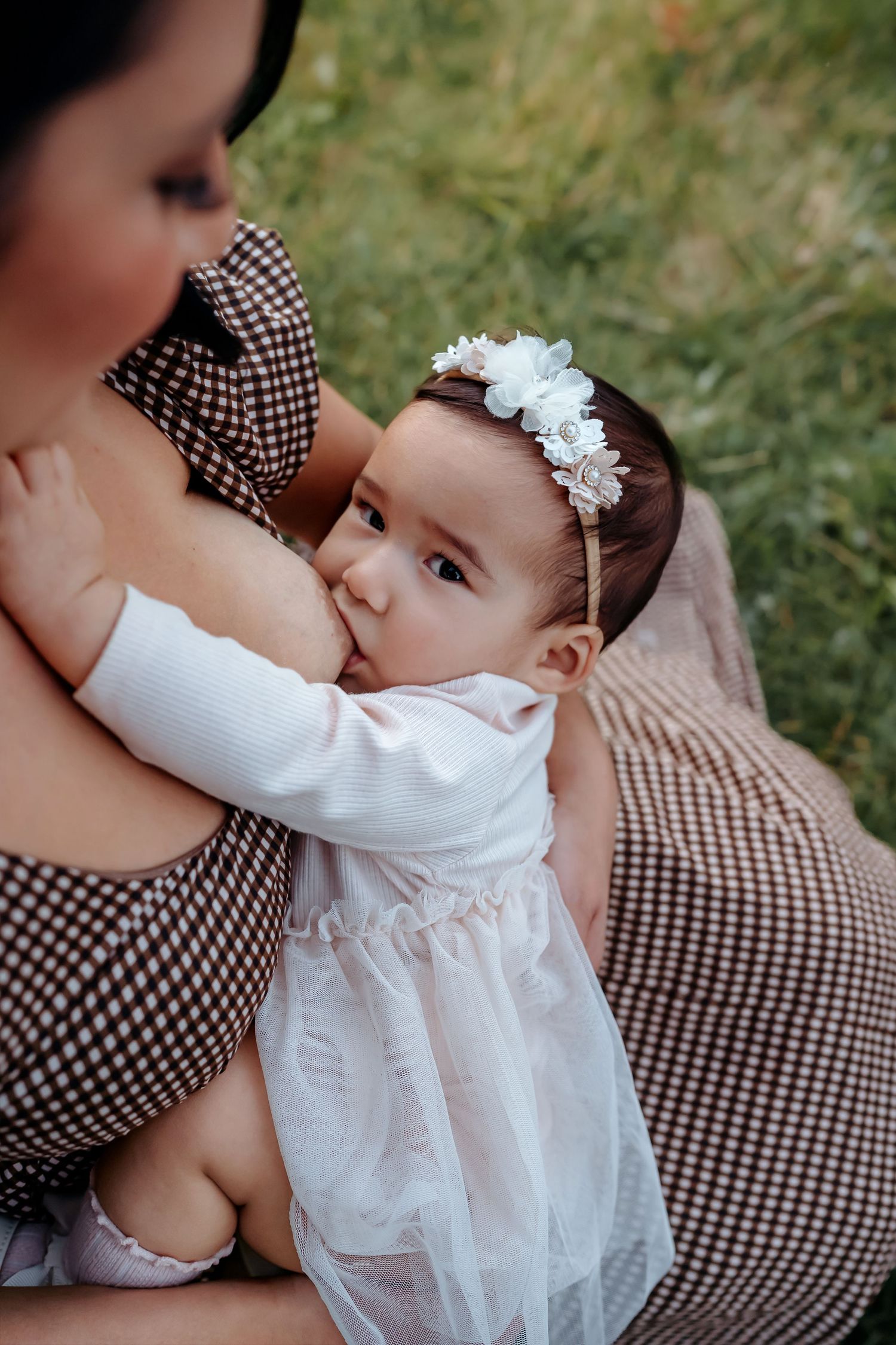 Baby in white dress rests against parent wearing checkered pattern dress in outdoor setting.