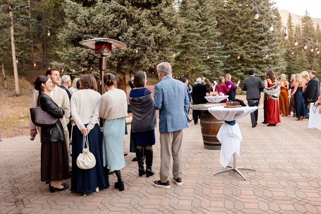 Wedding guests gather around a rustic wine barrel table during an outdoor evening reception with string lights overhead.