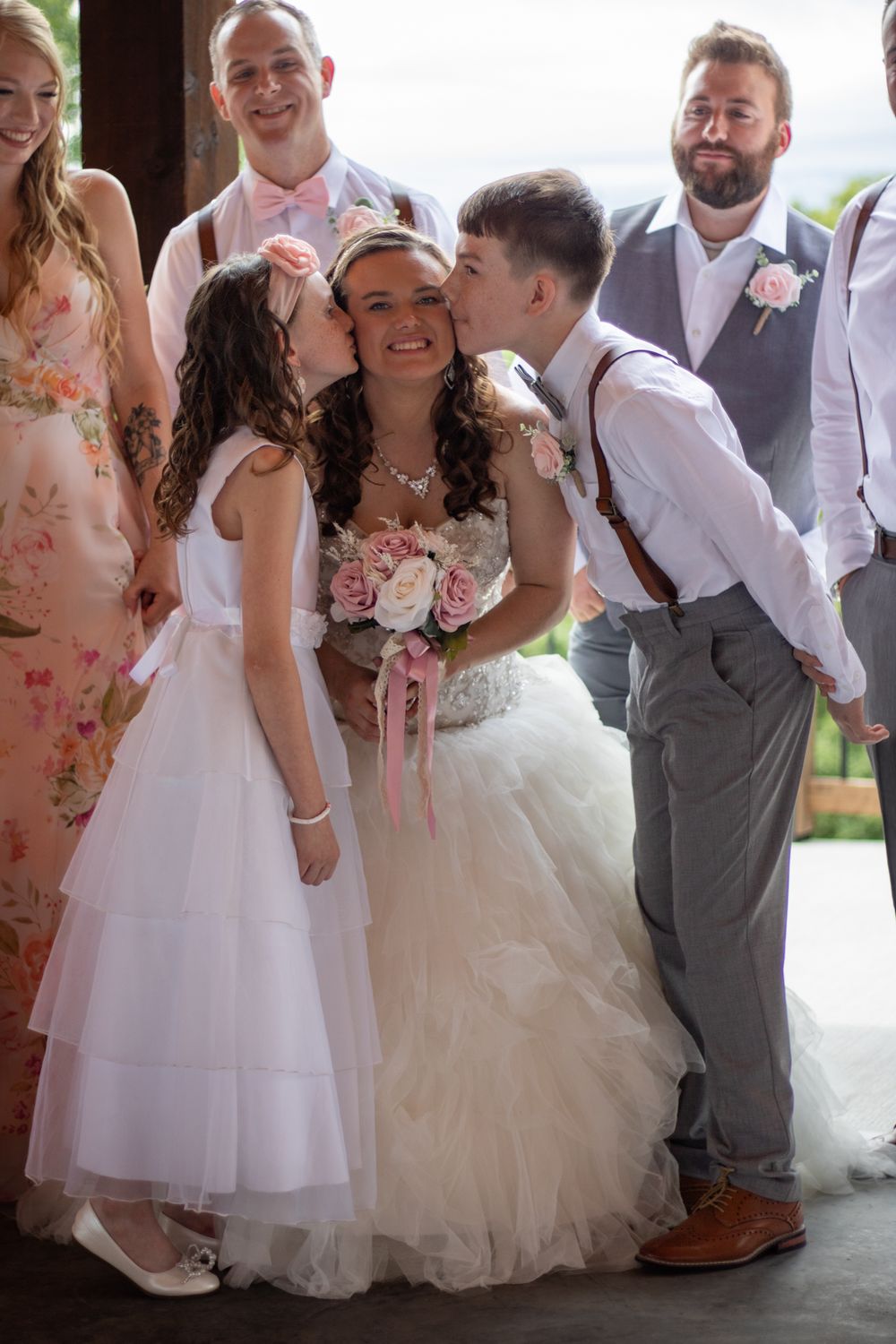 Flower girl and ring bearer kissing the brides' cheeks with bridal party behind them.