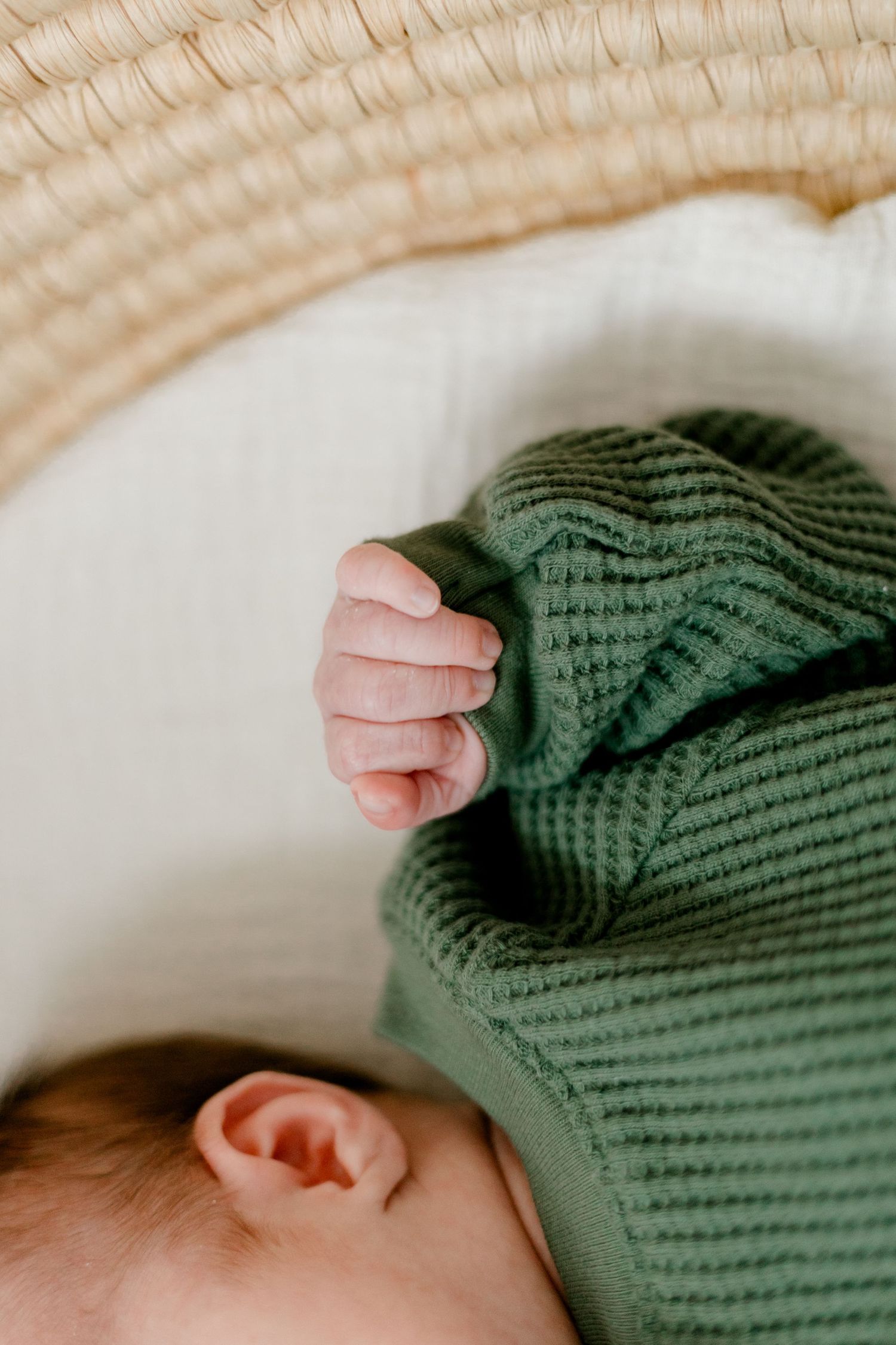 Newborn baby sleeping in a wicker bassinet wearing a green waffle knit outfit, tiny fist raised near ear.