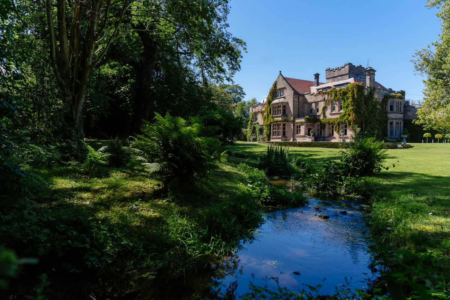 Scenic estate grounds with a tranquil pond reflecting trees and historic architecture. Ellingham Hall