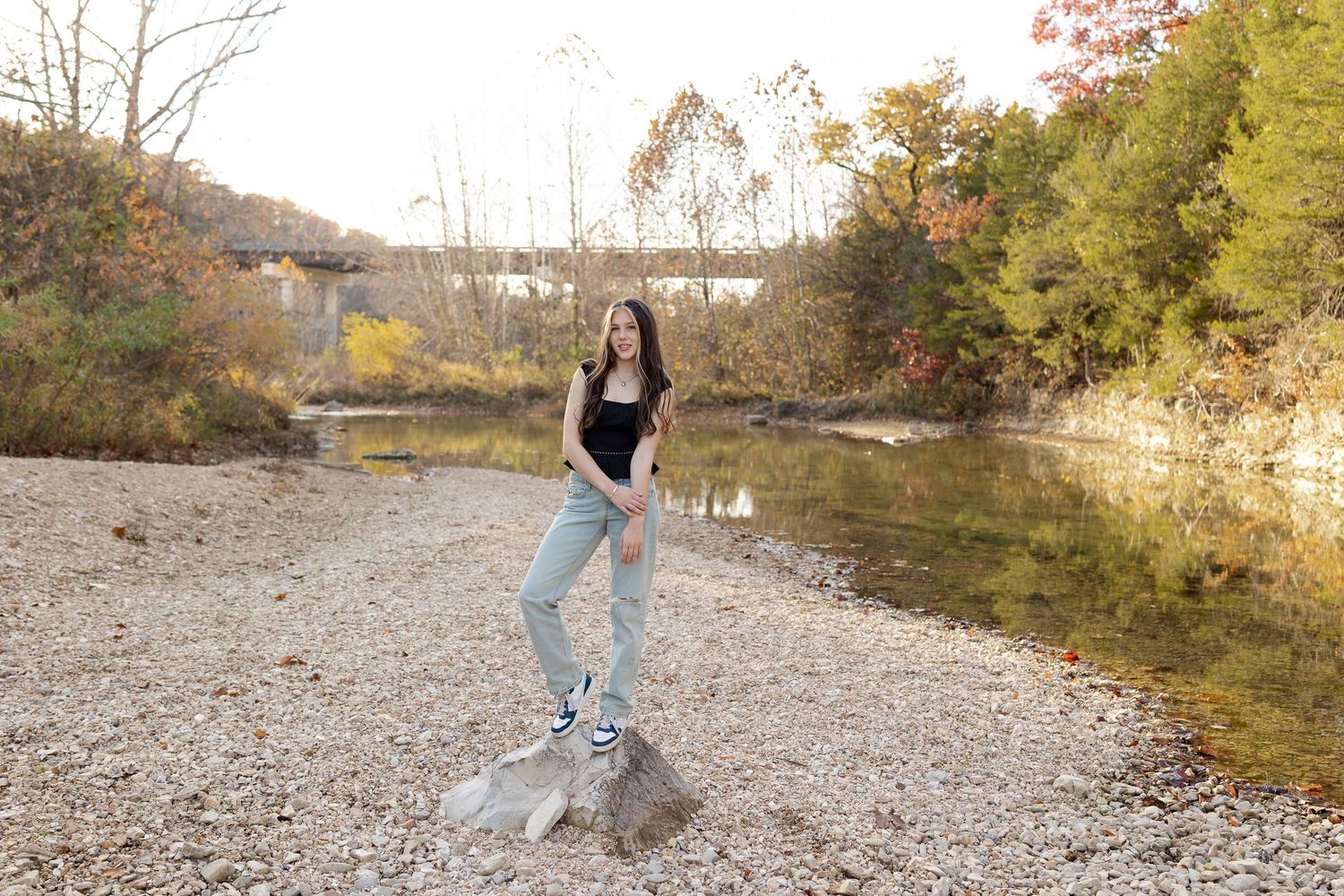 A person in jeans dances and jumps gracefully on a rocky riverbank surrounded by autumn foliage.