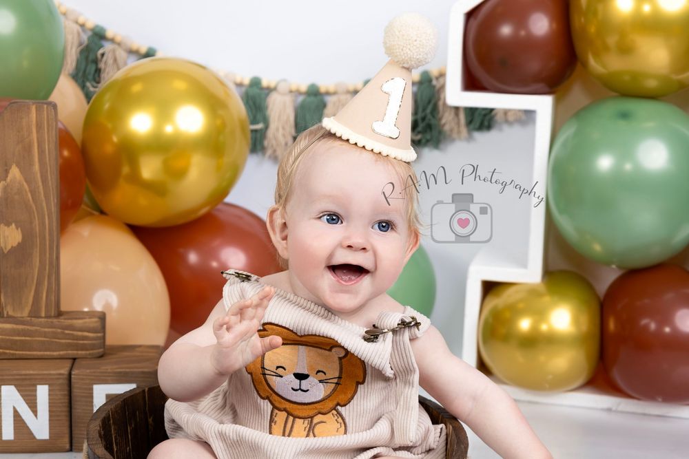 Baby smiling in lion outfit with party hat, surrounded by gold, green, and red balloons at a first birthday cake smash.R.A.M Photography