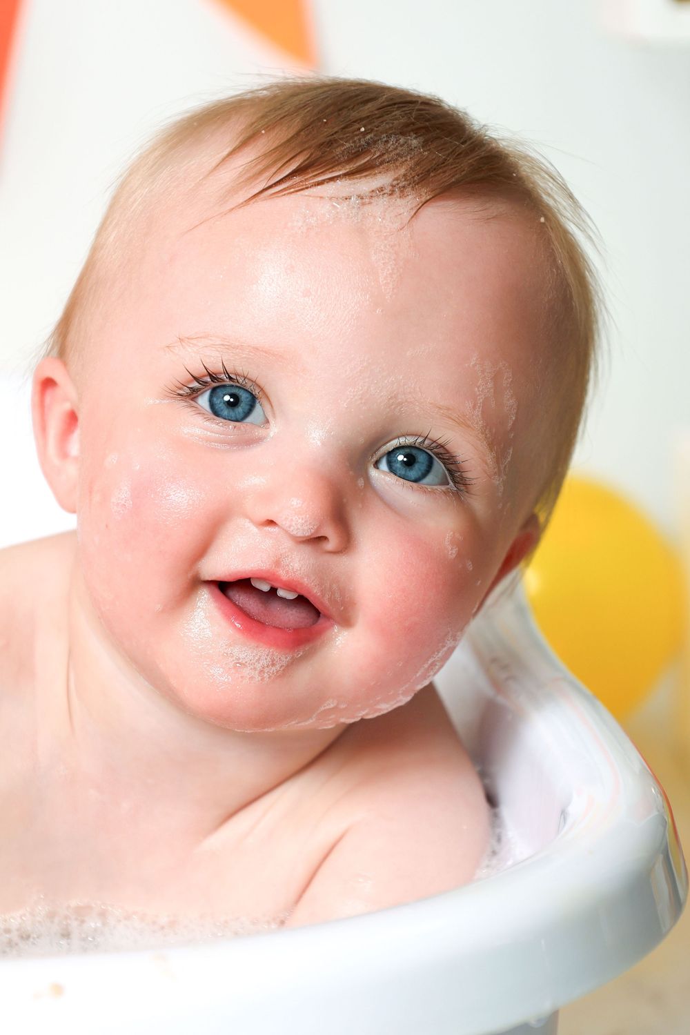 Adorable blue-eyed baby smiling during bath time with soap bubbles on face.