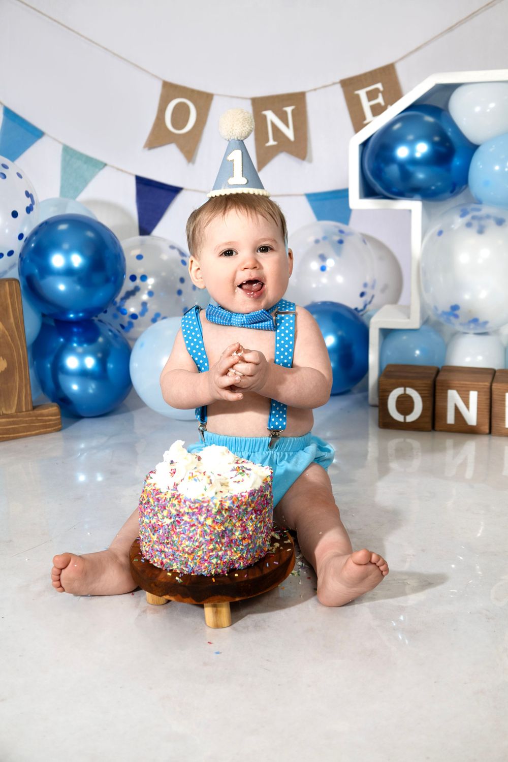 Baby boy in blue outfit smashes first birthday cake surrounded by blue balloons and ONE decorations.R.A.M Photography, Stranraer