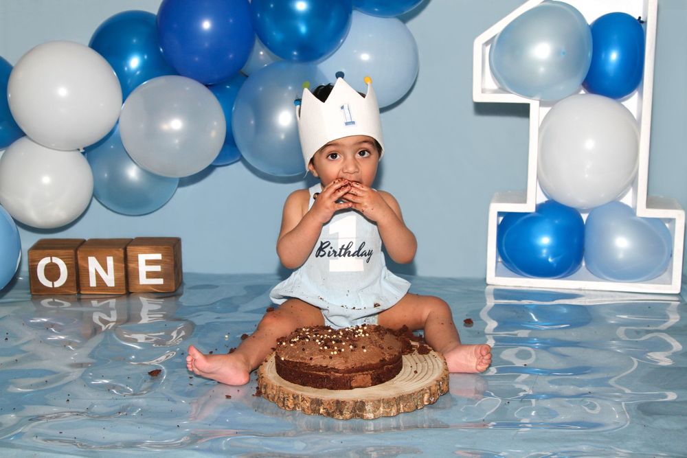 A cake smash photo session setup with blue and white balloons and a chocolate cake on a stand.