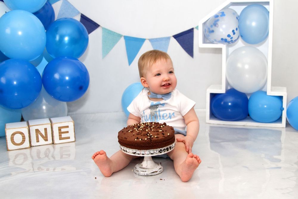 First birthday cake smash scene with blue balloons and 'ONE' blocks in the background.