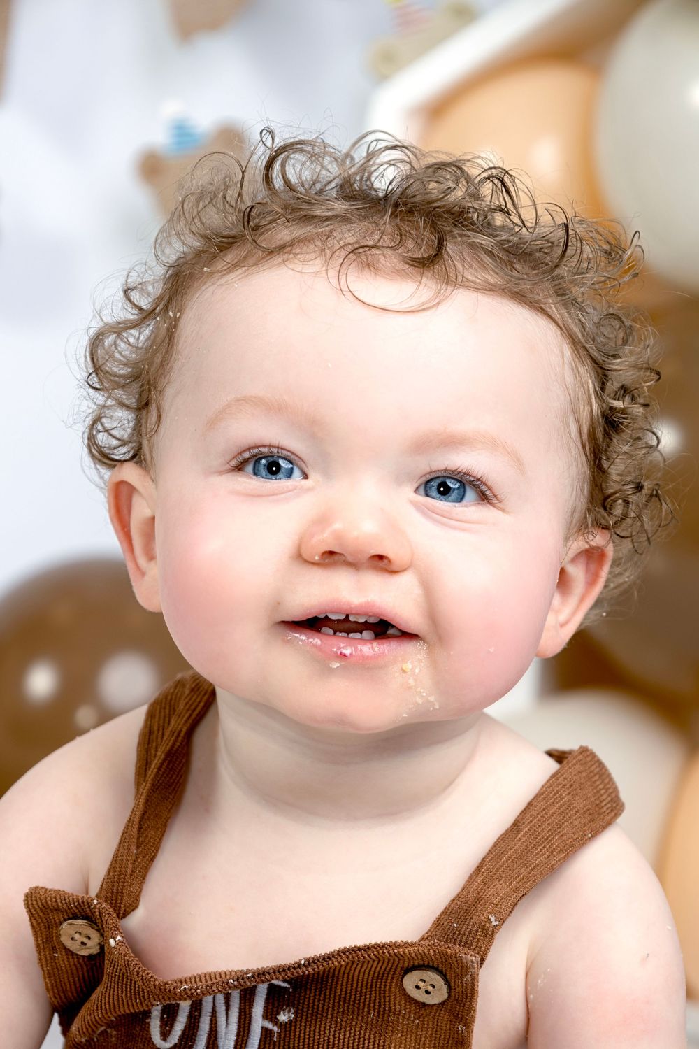 Adorable baby with blue eyes and curly hair wearing brown overalls smiles with cake on face at first birthday party.
