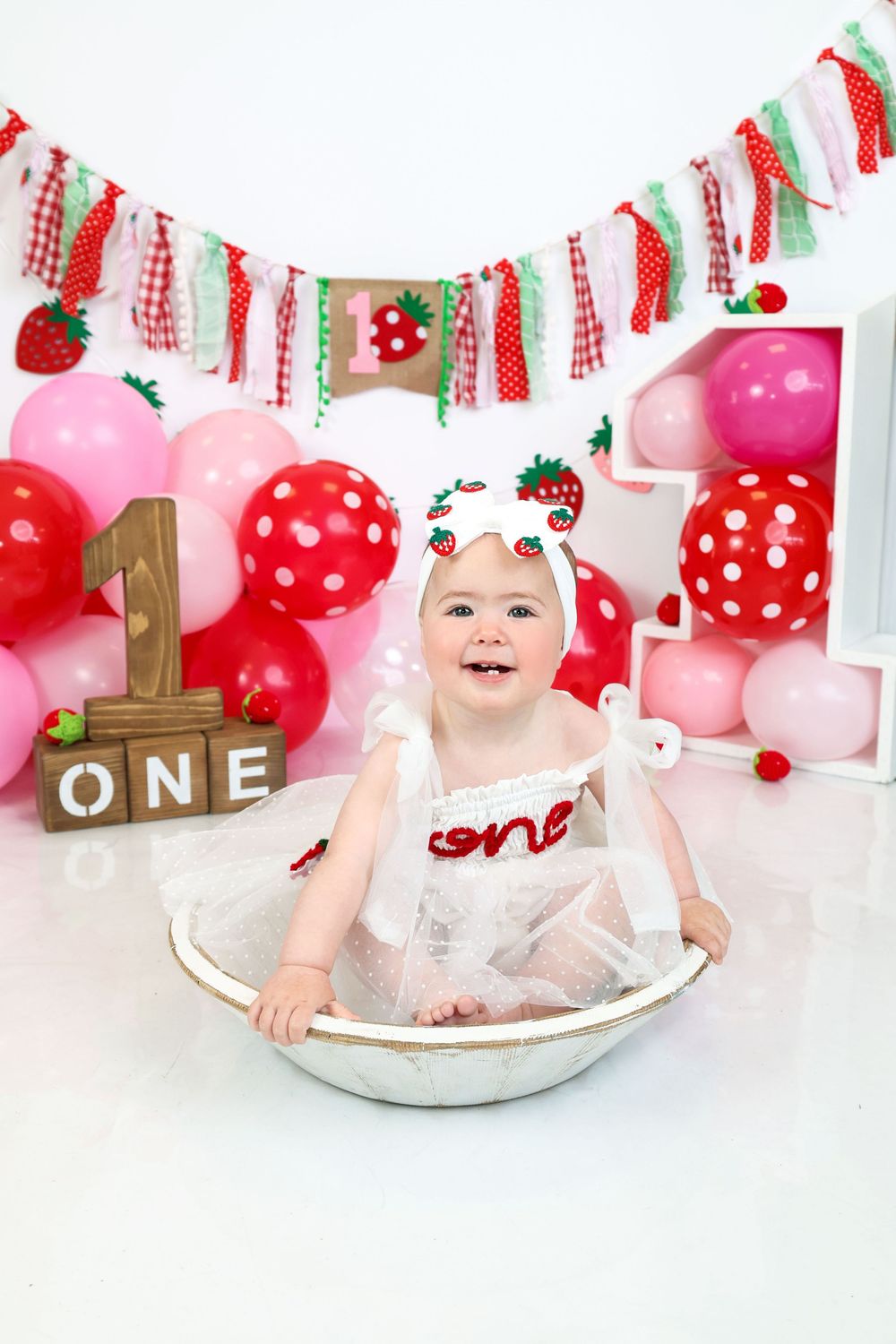 Baby girl in white strawberry outfit sits in a tin tub at a first birthday cake smash with red and pink balloon decor.R.A.M Photography,