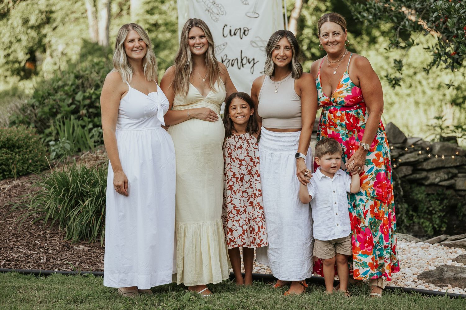 A group of people wearing summer dresses and floral prints pose together outdoors in a garden setting.