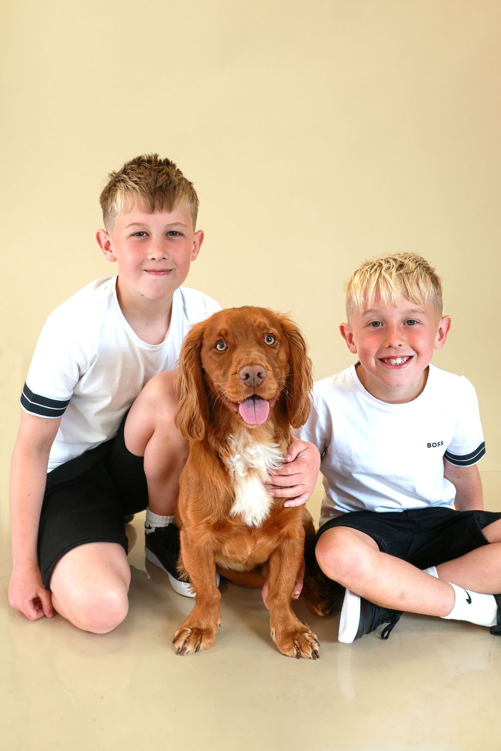 A brown Cocker Spaniel sitting between two children wearing matching white shirts against a beige background.