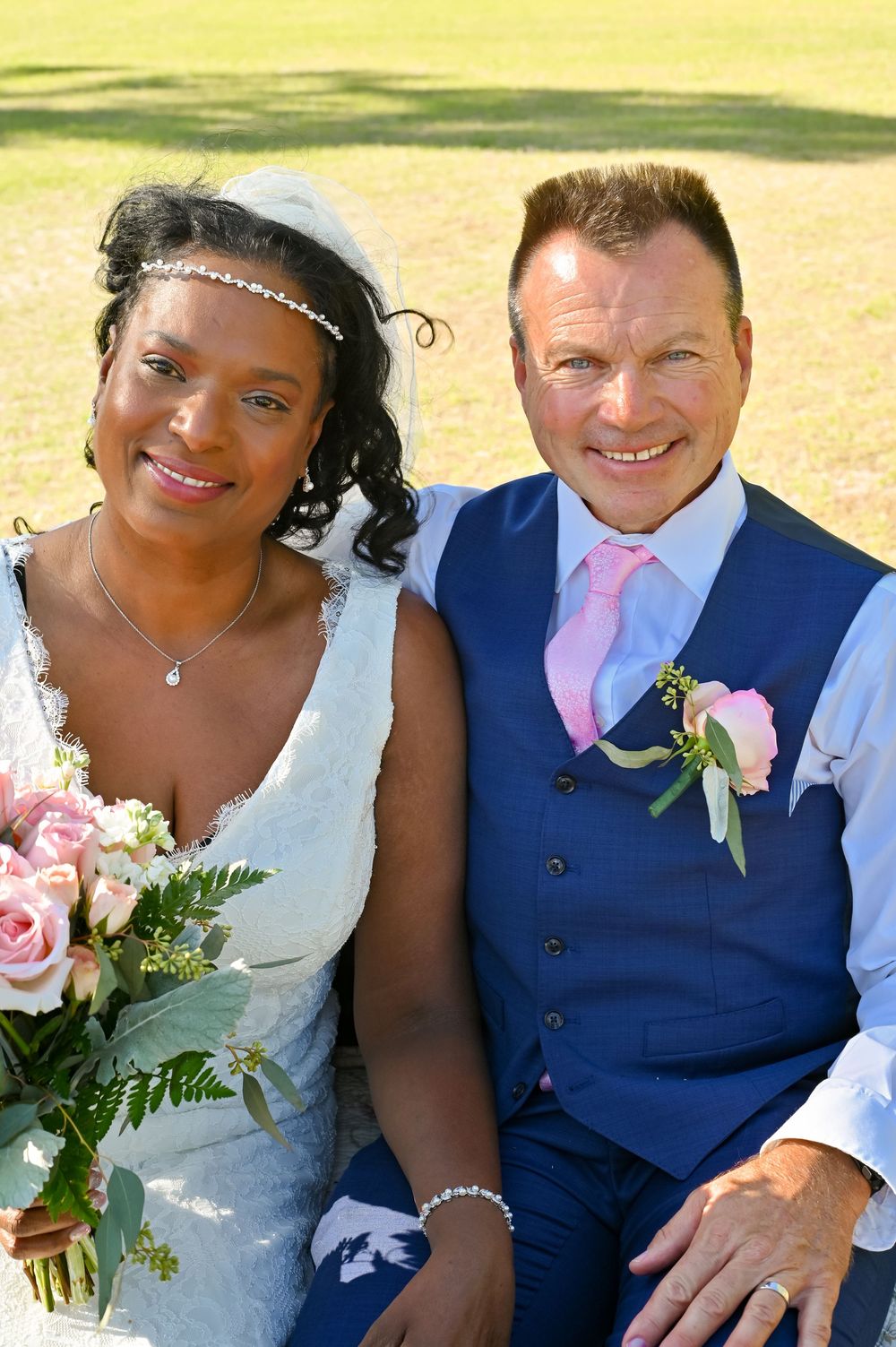 Happy newlywed couple in blue suit and white dress pose together outdoors with pink floral bouquet details.