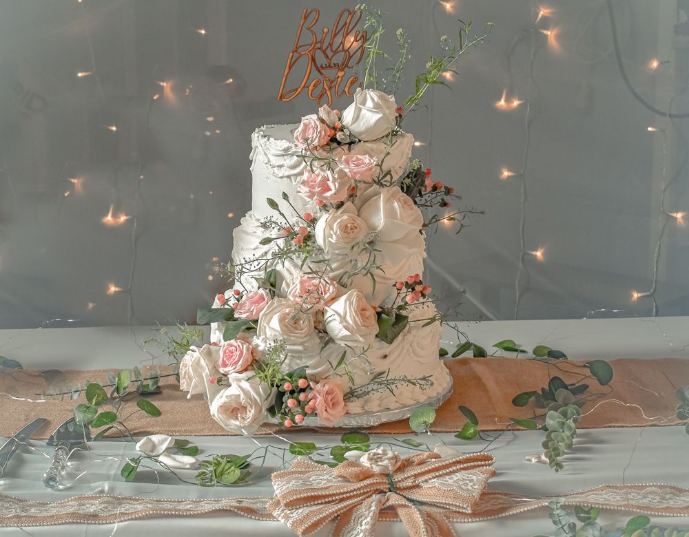 Elegant three-tier white wedding cake decorated with blush roses, eucalyptus, and a rose gold topper against twinkle lights.