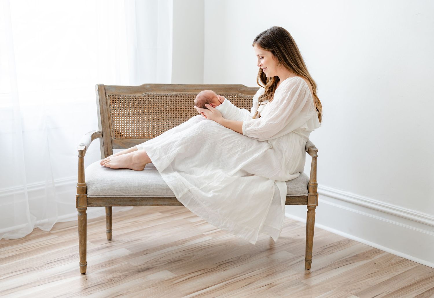 A person in a white dress sits barefoot on an elegant vintage-style settee against a light wall.