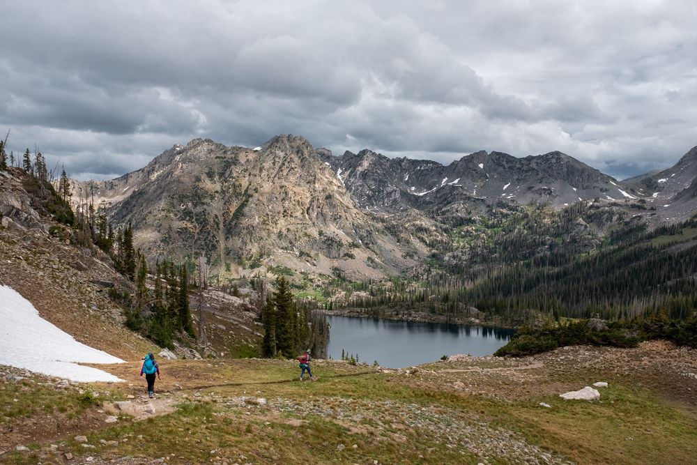 A hiker stands near a snowy patch overlooking a mountain lake surrounded by rugged peaks in the wilderness.
