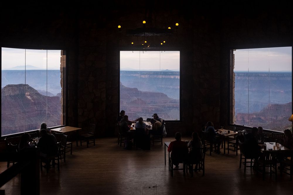 Restaurant dining room with panoramic windows overlooking mountain vistas at sunset, with empty tables silhouetted against the view.