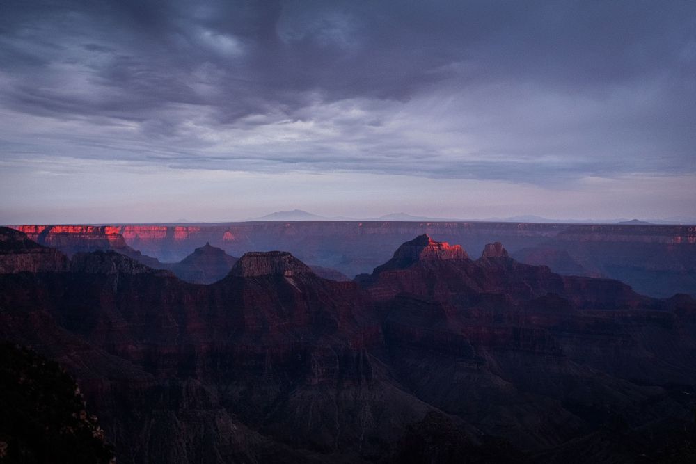 Panoramic view of the Grand Canyon at sunset with dramatic clouds and red-hued cliffs stretching to the horizon.