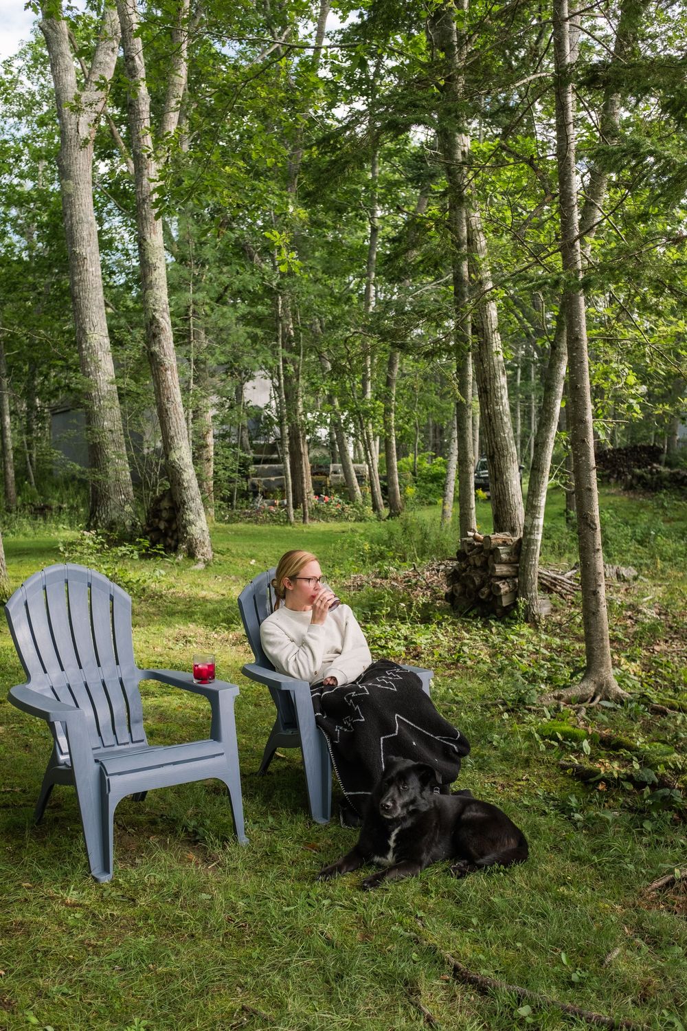 A sequence of blue Adirondack chairs in a wooded area with someone sitting and a black dog visible in the scene.