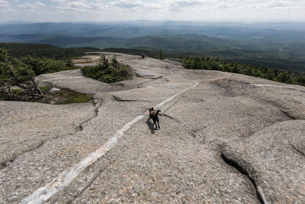 Hiker walking across exposed granite summit of mountain with sweeping views of forested valleys below.