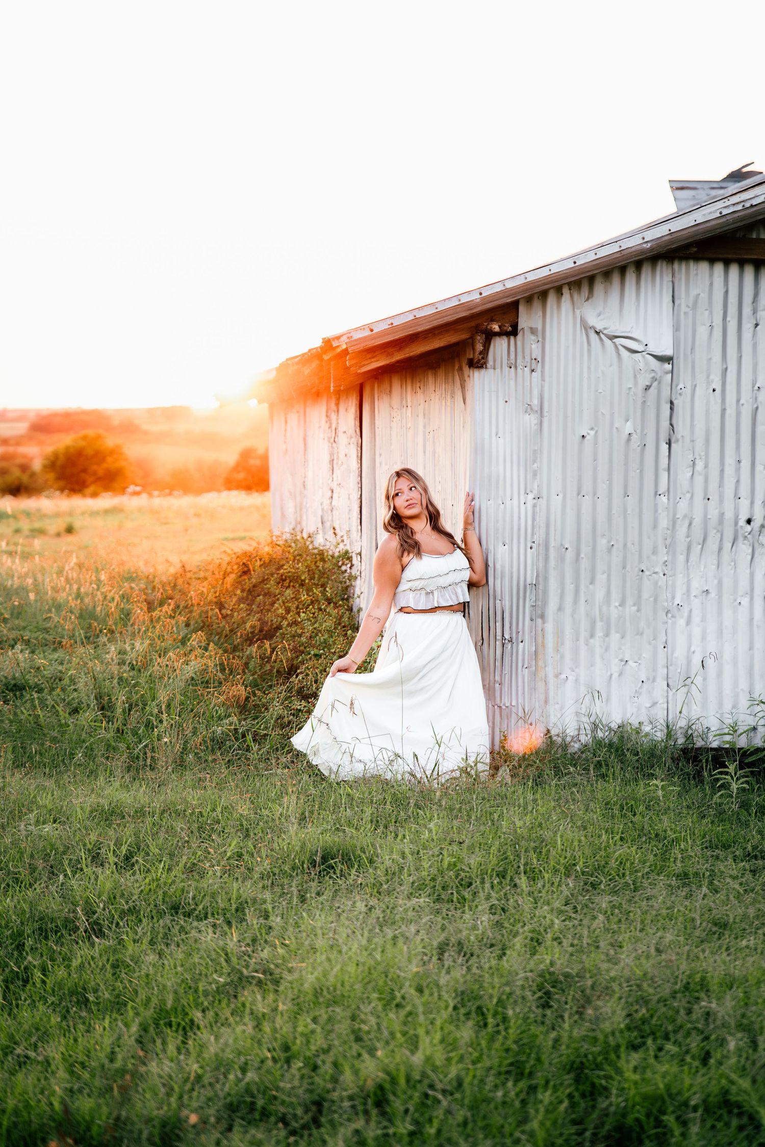 Woman in flowing white dress posing near rustic barn at golden sunset.