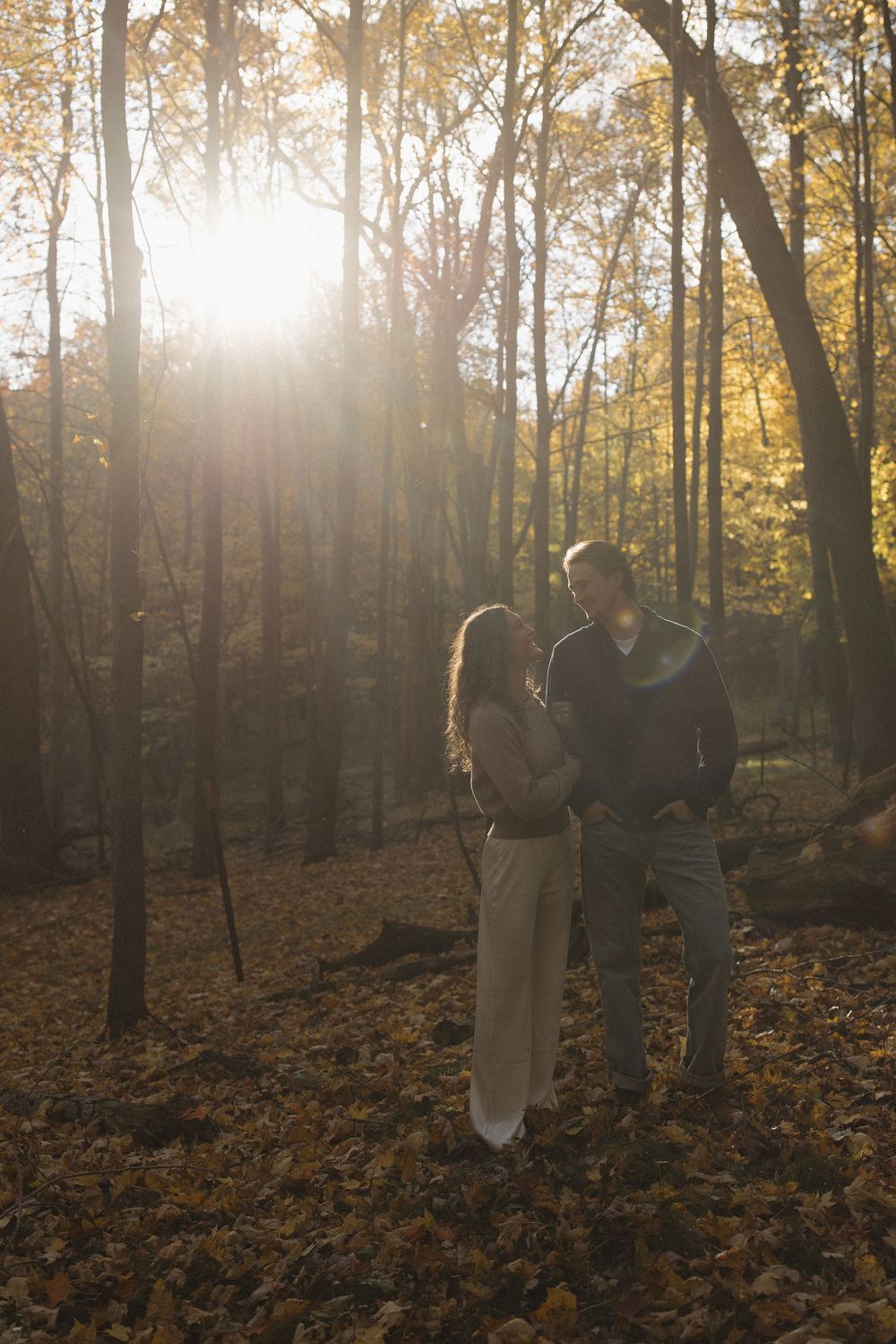 A couple stands close together in a sunlit autumn forest with golden leaves.