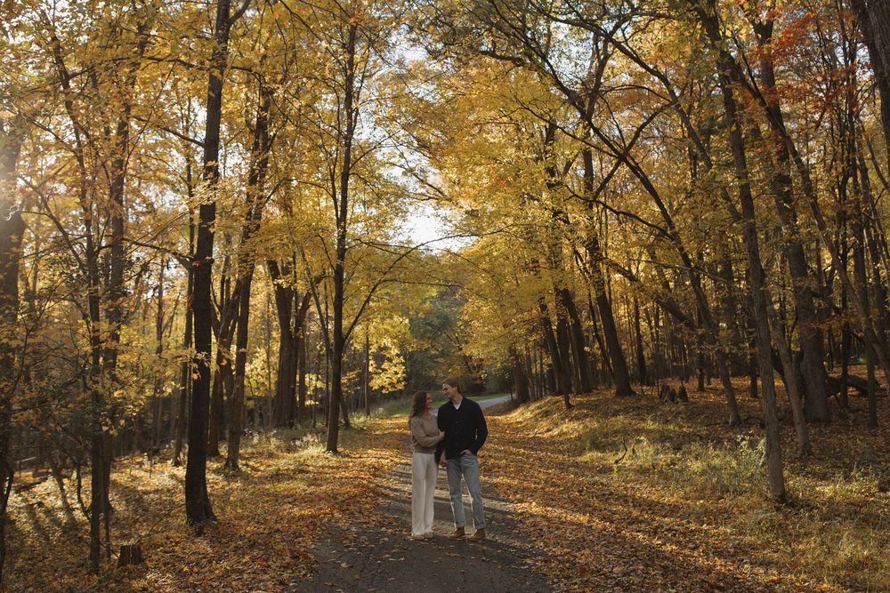 A couple walking together on a tree-lined path covered in fallen autumn leaves.