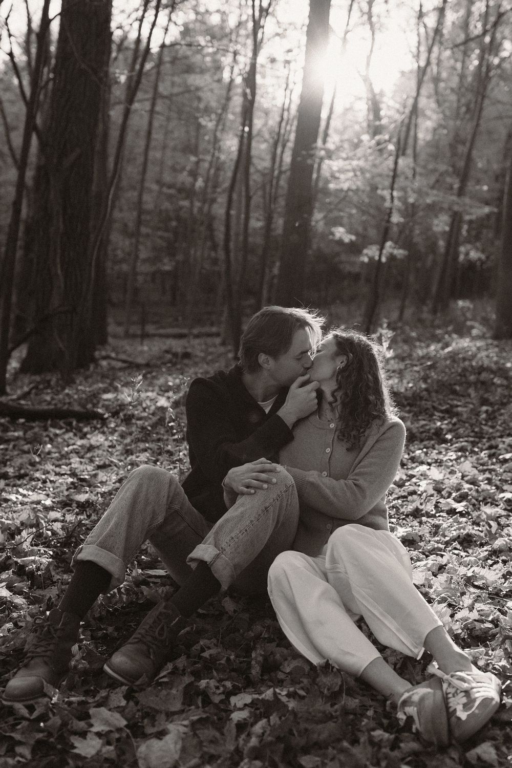 Black and white photo of a couple sharing an intimate moment among fallen leaves in the woods.