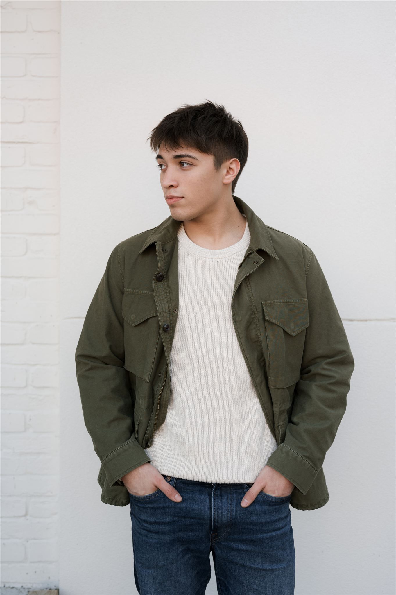 A person wearing an olive green utility jacket and white t-shirt poses against a white brick wall in a casual photoshoot.