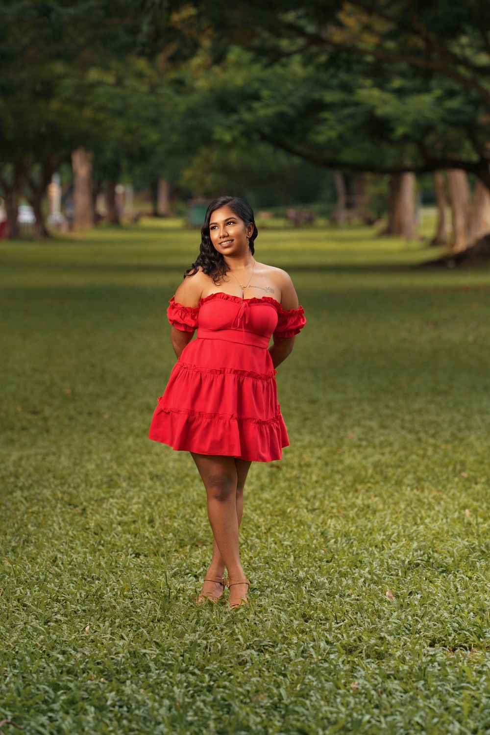 A person in a vibrant red off-shoulder dress poses gracefully on a grassy lawn with tall trees in the background.