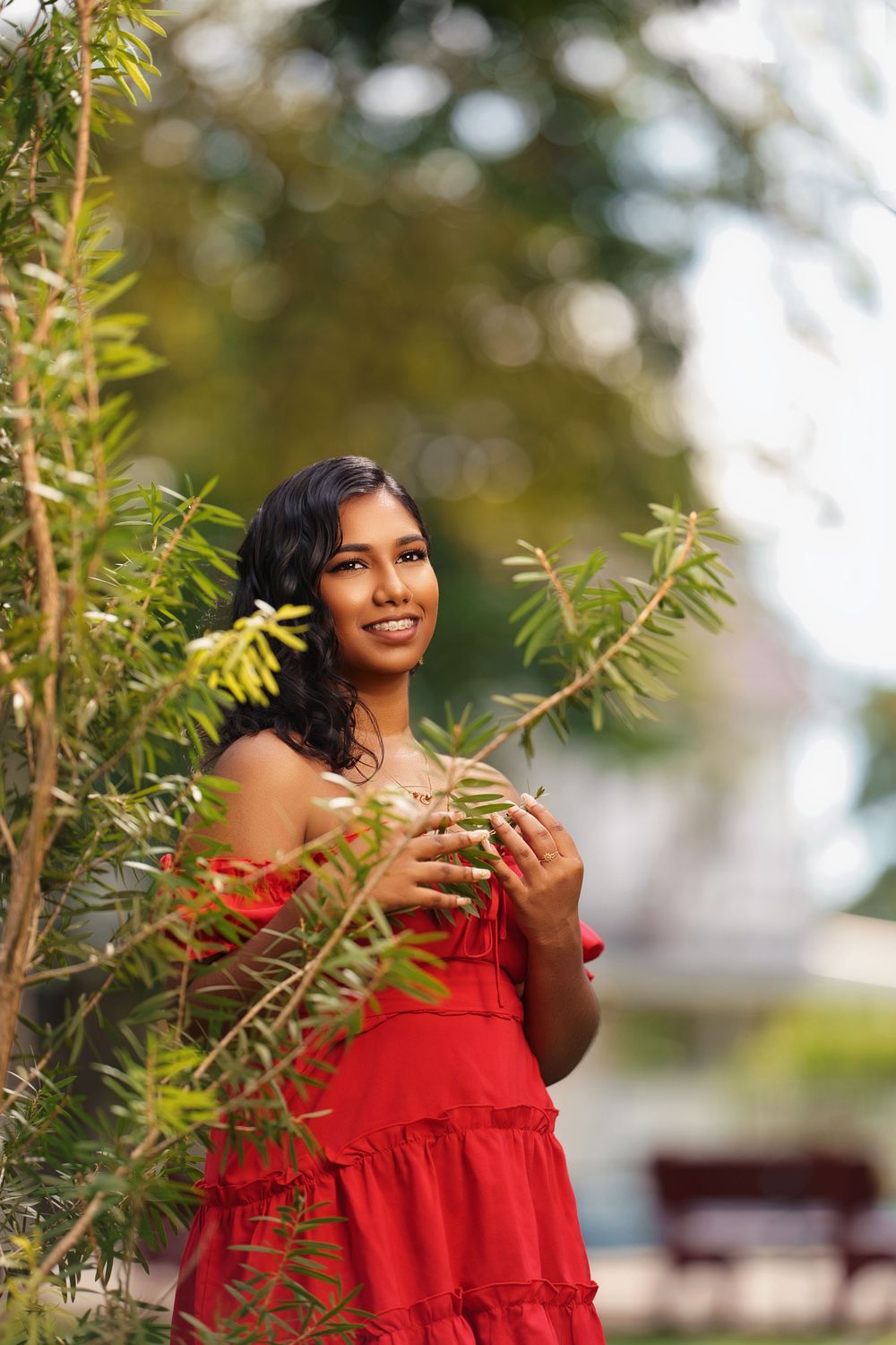 A woman in a vibrant red off-shoulder dress poses gracefully among green foliage in an outdoor setting.