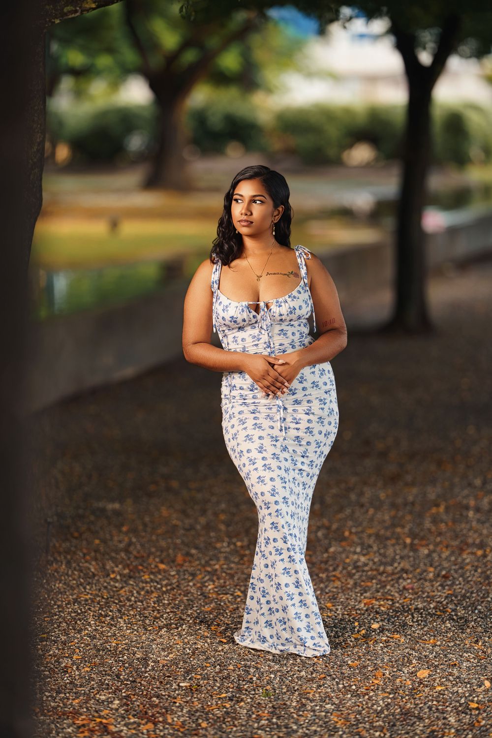 A white floral maxi dress is photographed on a path covered with fallen leaves under shady trees.