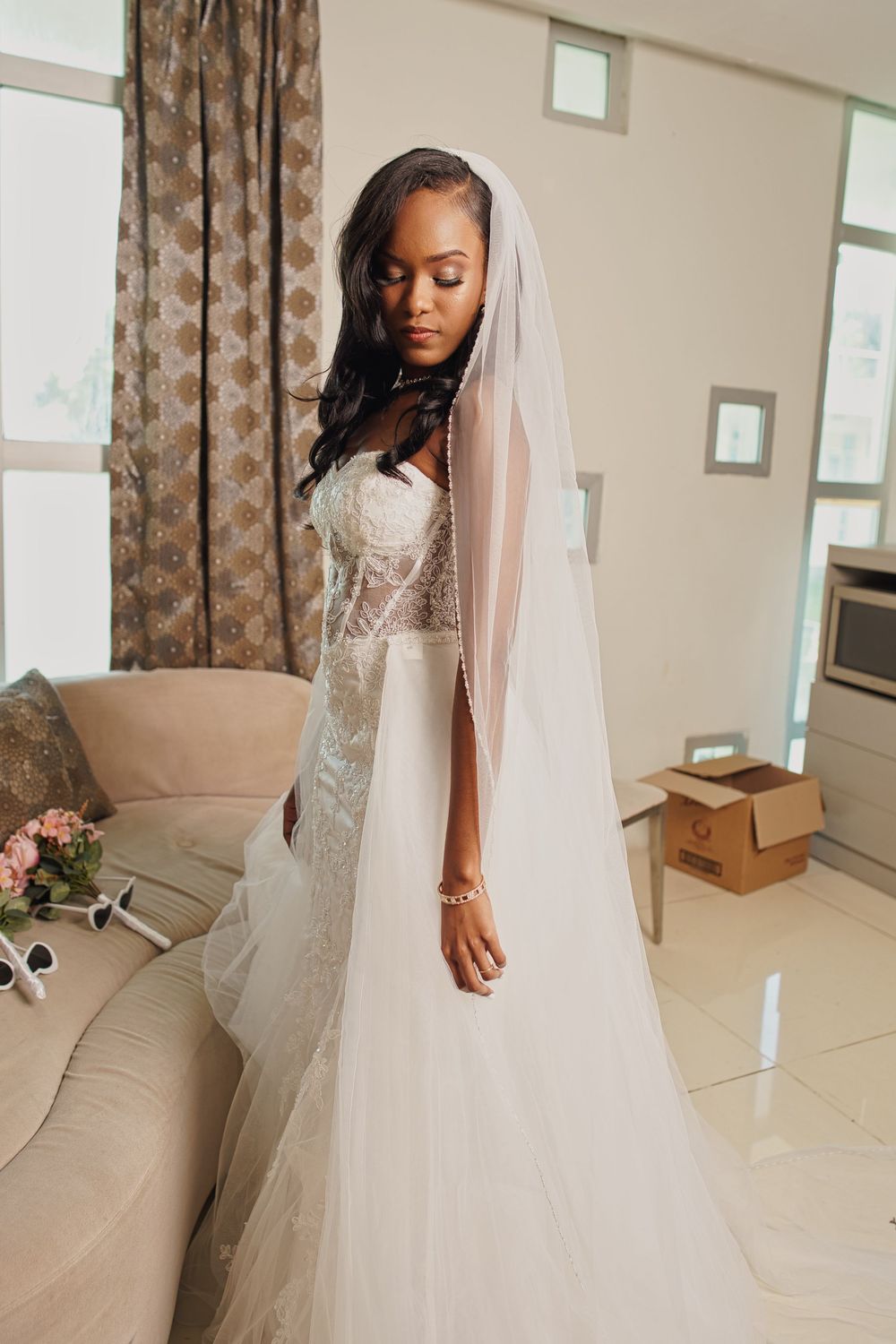 A bride in an elegant white lace wedding gown poses gracefully in a sunlit room with flowing veil and floral details.
