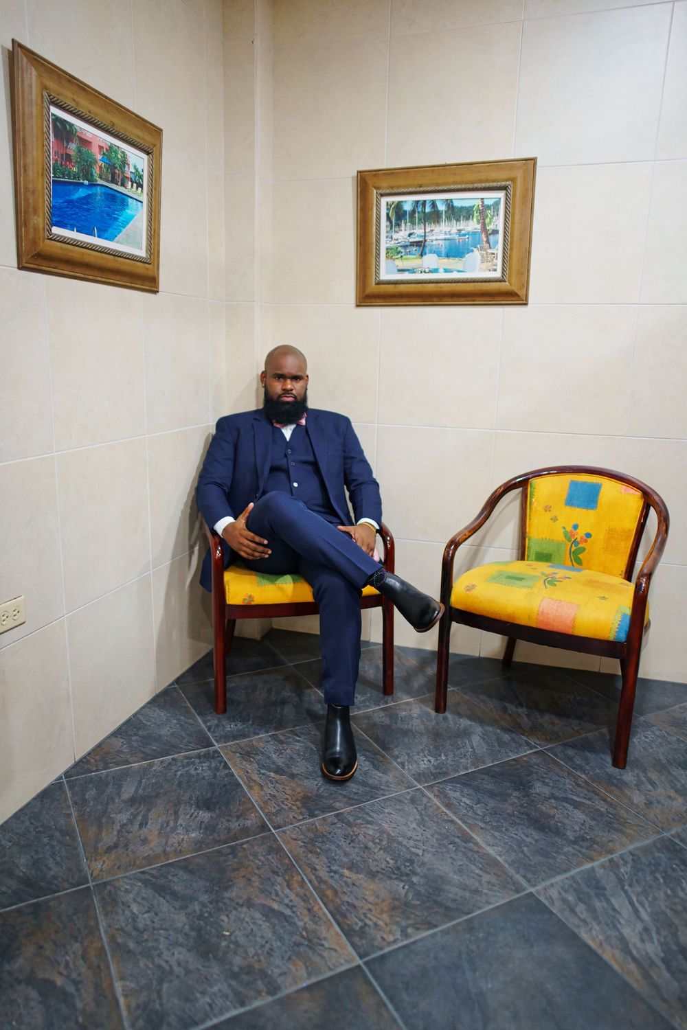 A groom in a navy blue suit sits casually on a bench in a hallway decorated with framed artwork.
