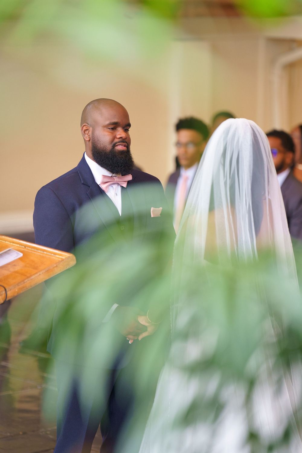 Wedding ceremony guests in formal attire gather inside an elegant venue with soft lighting and greenery decor.