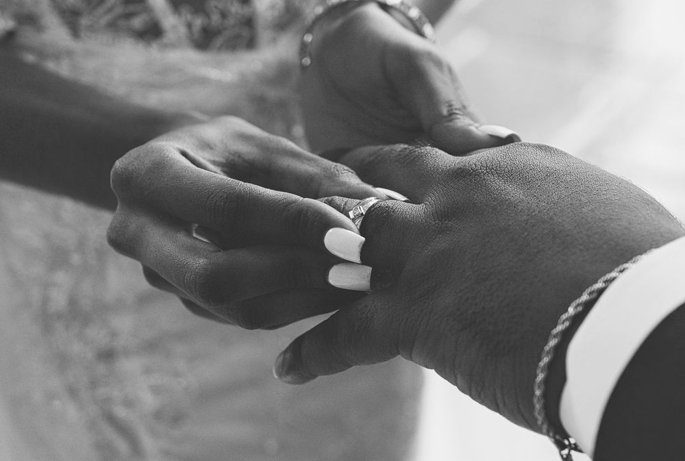 A close-up black and white photo shows hands exchanging a diamond engagement ring during a proposal moment.