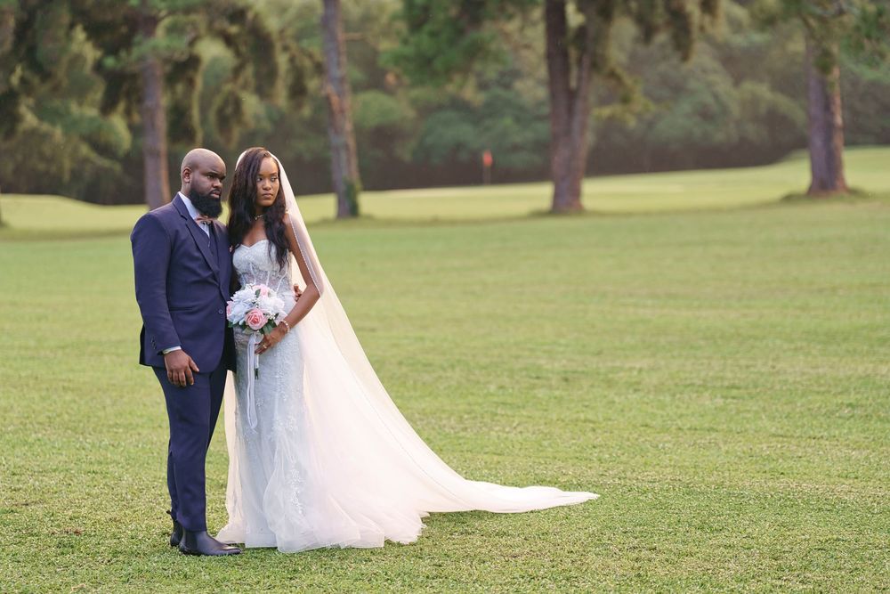 A newlywed couple stands together on a lush green lawn surrounded by tall pine trees in an outdoor setting.