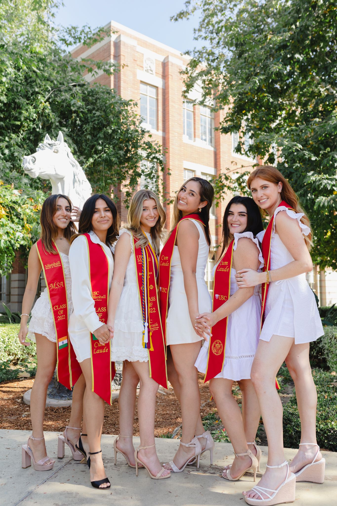 A group of college graduates in white dresses and red sashes pose together on a sunny day at the University of Southern California.