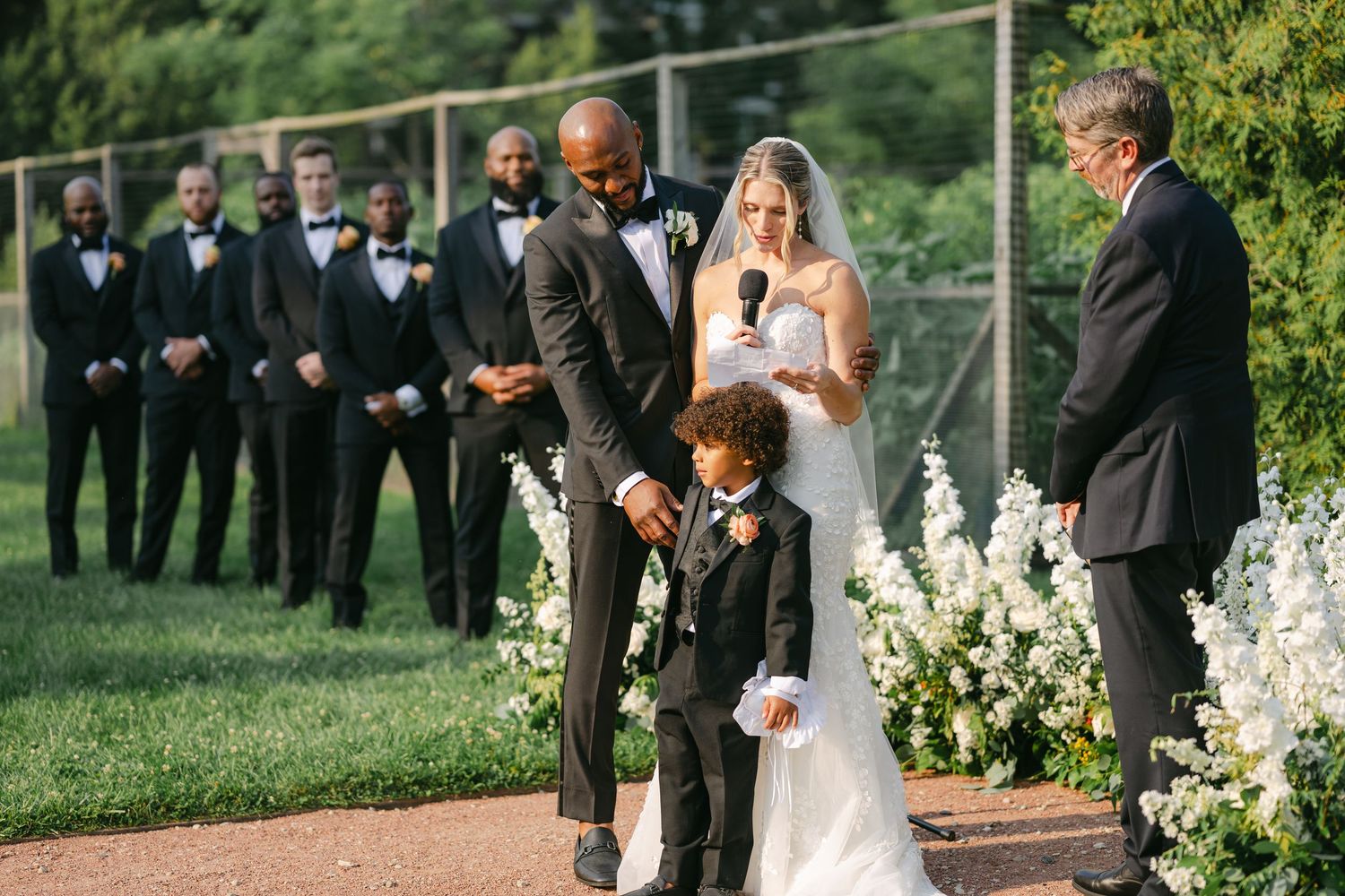 Outdoor wedding ceremony setup with white floral arrangements and guests in formal attire gathered on a green lawn. - Elawa Farm