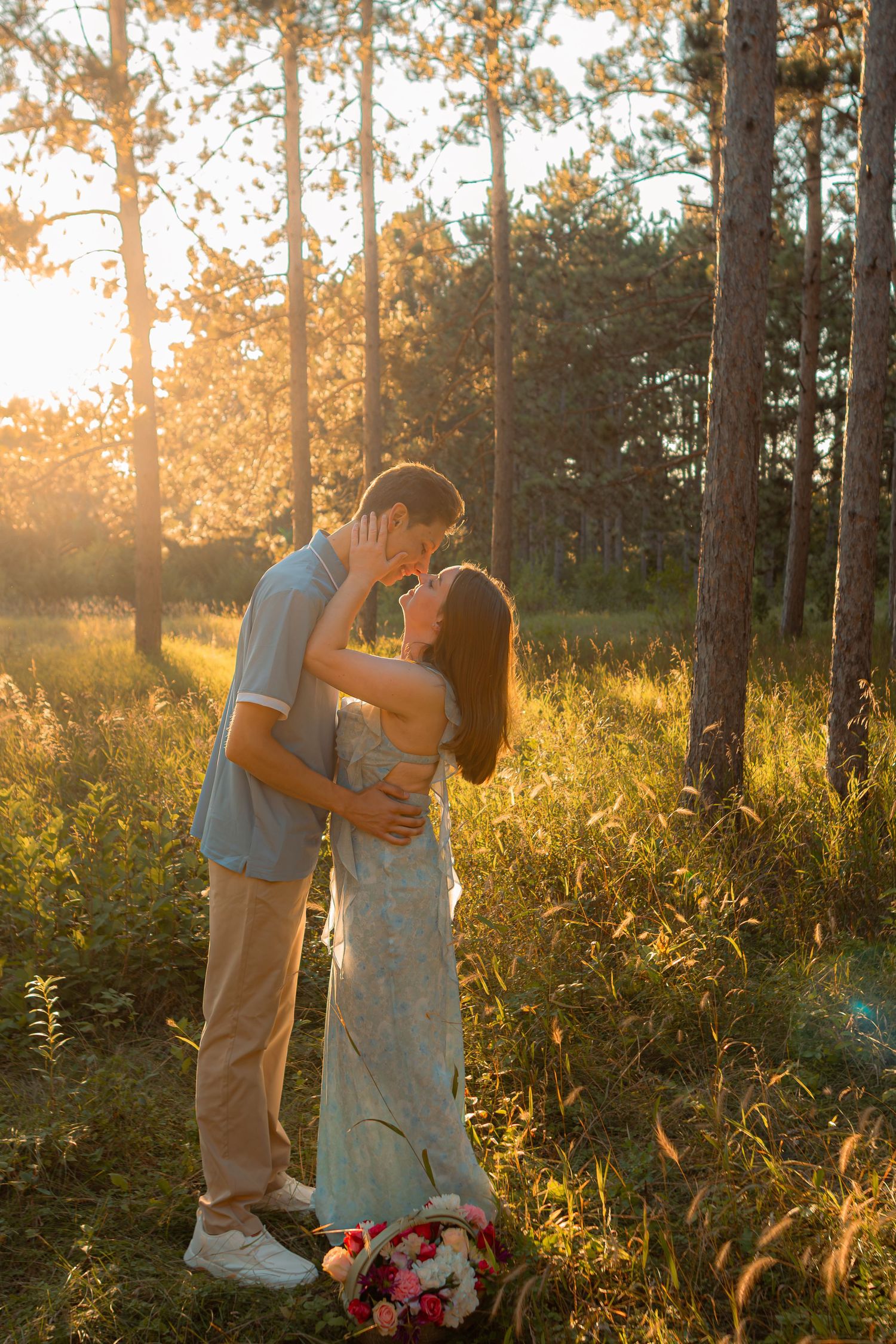 Couple sharing a romantic kiss in a sunlit forest clearing, with golden hour light and a floral bouquet at their feet.
